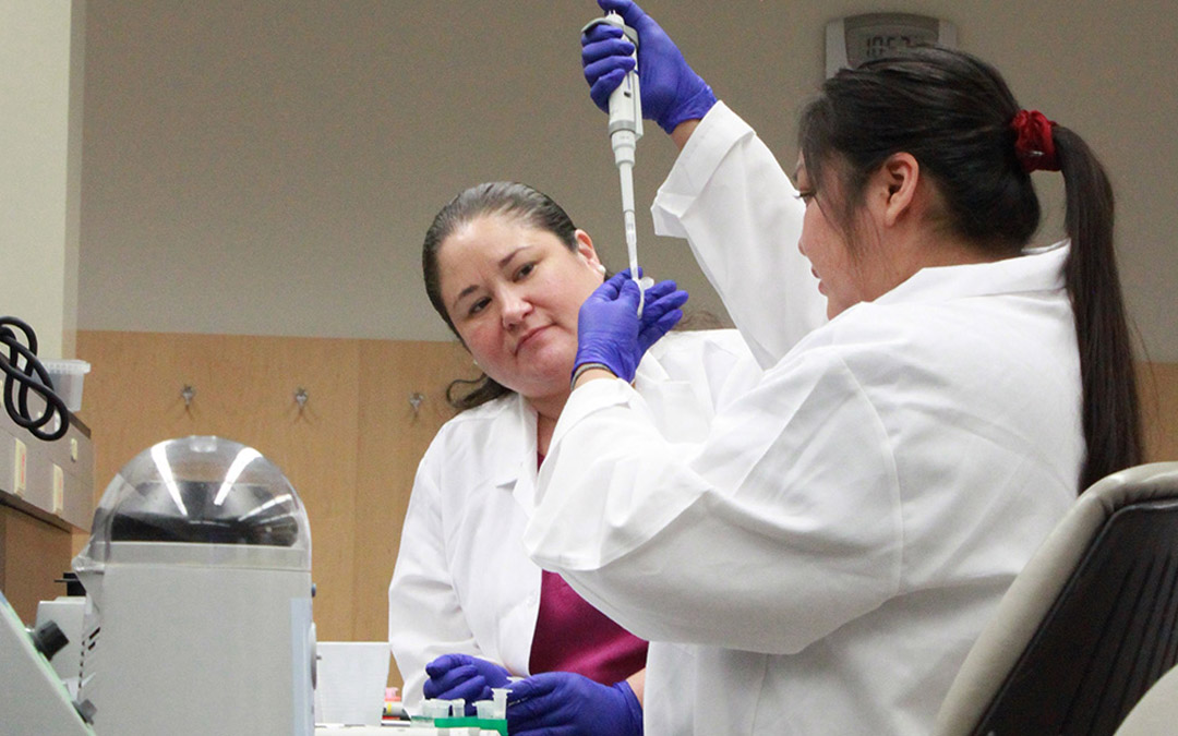 Johns Hopkins post-doctoral researcher Dr. Jessica L. Elm, citizen of the Oneida Nation and descendant of the Stockbridge-Munsee Band of the Mohicans, watches Alison Watson of the Navajo Nation as she receives hands-on training in genomics research at the 2019 SING workshop.